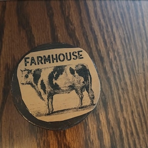 May include: A circular dark brown wooden object featuring a tan paper label on top. The label displays a black ink illustration of a cow standing in profile, with the word FARMHOUSE printed in bold black capital letters above the animal.