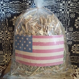 May include: A clear plastic bag filled with dried hay, with a vintage American flag sticker on the front. The bag is sitting on a wooden surface.