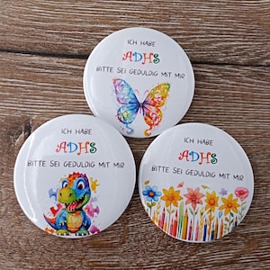 Può includere: Tre bottoni rotondi bianchi con disegni colorati. Ogni bottone ha il testo "Ich habe ADHS Bitte sei geduldig mit mir" che si traduce in "Ho ADHD Per favore, sii paziente con me". I disegni includono una farfalla, un dinosauro e fiori e matite colorate.