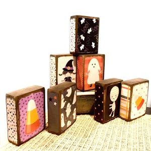May include: A collection of Halloween-themed wooden blocks. Each block features a different design, including a witch, ghost, candy corn, and patterned backgrounds. The blocks are painted with a dark brown border.
