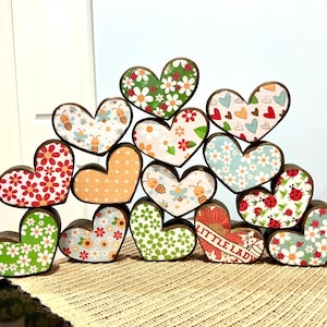 May include: A collection of heart-shaped wooden blocks featuring various patterns including daisies, ladybugs, bees, and hearts. One heart displays the text LITTLE LADY in red lettering. The blocks are arranged in a stacked, pyramid-like formation.