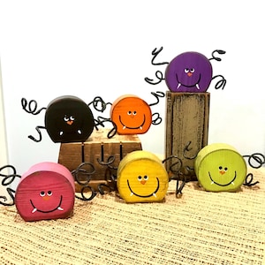 May include: A collection of colorful, wooden spider decorations with smiling faces and wire legs. The spiders are painted in various colors including black, orange, purple, pink, yellow, and green. Perfect for Halloween.