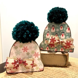 May include: Two decorative wooden winter hat ornaments. Each hat features a teal pom-pom, snowflake patterned paper, and a burlap bow with red snowflake accents. One hat is on top of a wooden block.