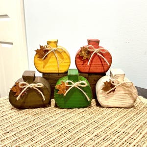 May include: Five wooden pumpkin-shaped decorations in yellow, red, brown, green, and white. Each pumpkin features a small brown artificial leaf and a tied jute twine bow around the stem. The pumpkins have a ribbed texture and a flat base.
