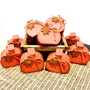 May include: A collection of orange wooden pumpkin-shaped decorations, each featuring a brown stem, a tied piece of orange twine, and a small metallic leaf accent. Several pumpkins are arranged inside a rectangular wooden crate.