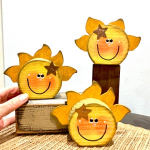 May include: Three yellow wooden sun decorations with painted smiling faces, rosy cheeks, and a small brown star accent on each. The suns feature jagged rays around the circular center and are finished with a distressed, rustic wood grain texture.