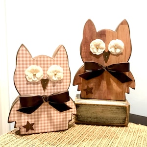 May include: Two wooden owl figurines. One is patterned with a pink and white gingham design, the other is a natural wood tone. Both have cream-colored flower eyes, brown bows, and star accents. One owl sits on a small, decorative block.