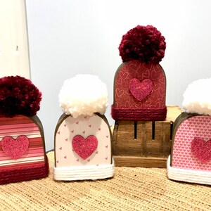 May include: Four wooden Valentine's Day decorations shaped like hats with pom-poms on top. Each hat has a different pink and white patterned design with a glittery pink heart in the center.