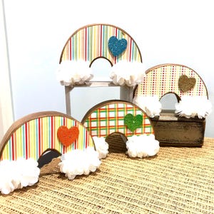 May include: Four wooden rainbow arches with different patterned paper, each with a heart-shaped embellishment. The hearts are different colors and some have glitter. Each arch has a white fluffy cloud underneath.