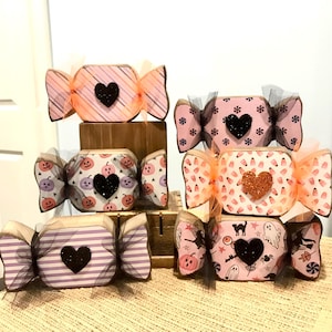 May include: A collection of decorative Halloween candy-shaped ornaments. Each ornament features a unique design, including stripes, pumpkins, and bats, with a glitter heart in the center. The ornaments are wrapped with sheer ribbon.