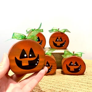 May include: A collection of orange wooden pumpkin decorations with black painted faces and green ribbon bows. The pumpkins have a smiling jack-o'-lantern design with white accents. These Halloween-themed decorations are perfect for seasonal home decor.