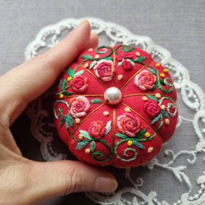 May include: A red pincushion with a pearl in the center, embroidered with pink and red roses, green leaves, and yellow flowers.