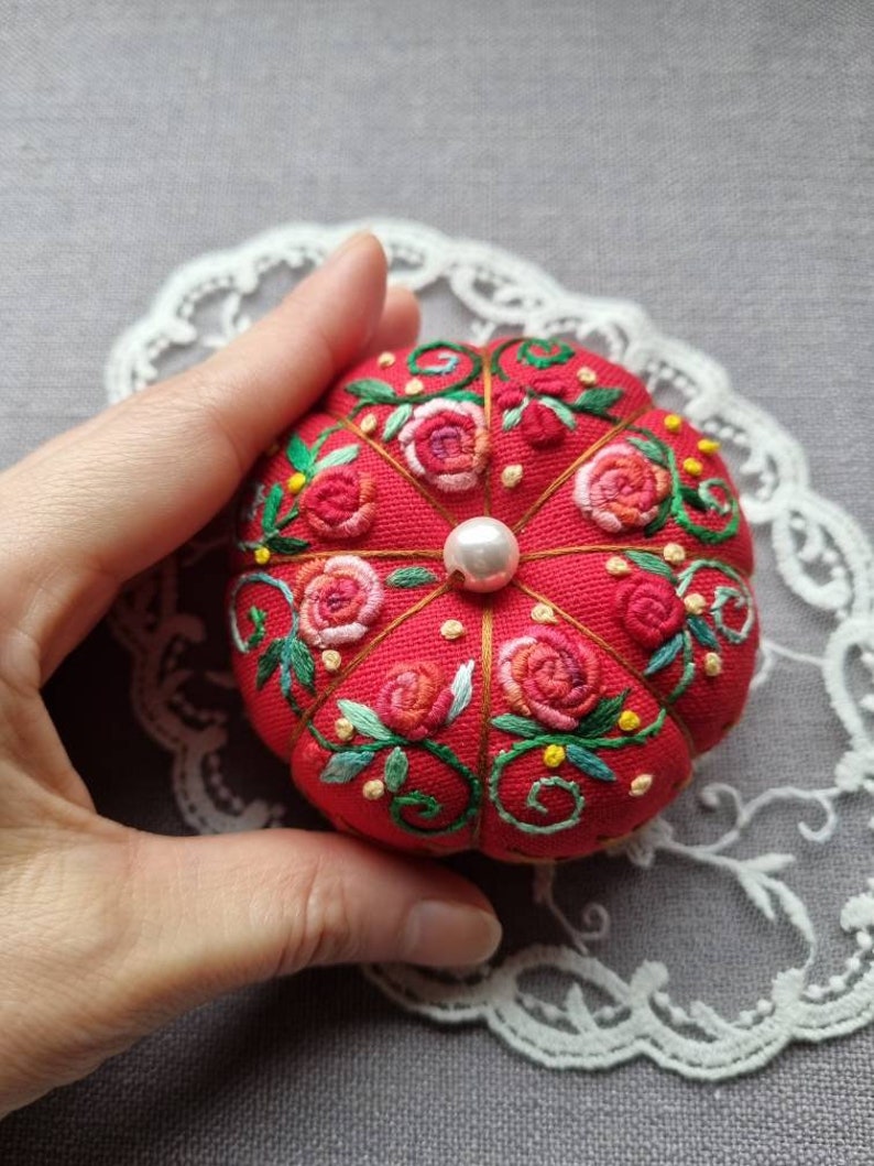 May include: A red pincushion with a pearl in the center, embroidered with pink and red roses, green leaves, and yellow flowers.