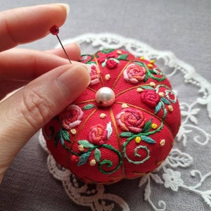 May include: A red pincushion with a pearl button in the center, surrounded by embroidered roses and vines. A hand is holding a pin with a red head over the pincushion.