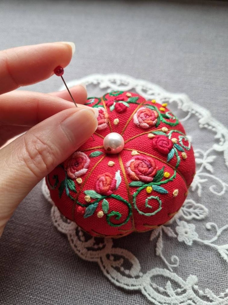 May include: A red pincushion with a pearl button in the center, surrounded by embroidered roses and vines. A hand is holding a pin with a red head over the pincushion.