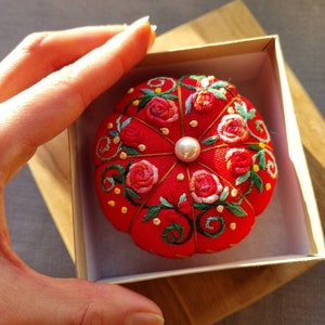 May include: A red pincushion with a pearl in the center, embroidered with pink and green flowers. The pincushion is in a white box with a wooden base.