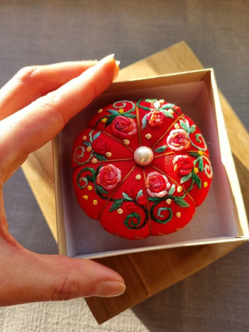 May include: A red pincushion with a pearl in the center, embroidered with pink and green flowers. The pincushion is in a white box with a wooden base.