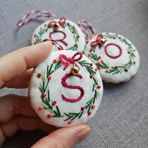 Peut inclure: Trois ornements ronds en tissu blanc avec des couronnes brodées à la main en vert, agrémentées de baies rouges et d'un nœud rouge. Chaque pièce présente une lettre rouge brodée au centre, une petite clochette dorée et une ficelle bicolore.