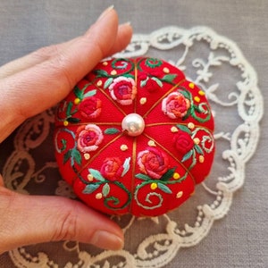 May include: A red pincushion with a pearl button in the center, embroidered with pink and red roses and green leaves.