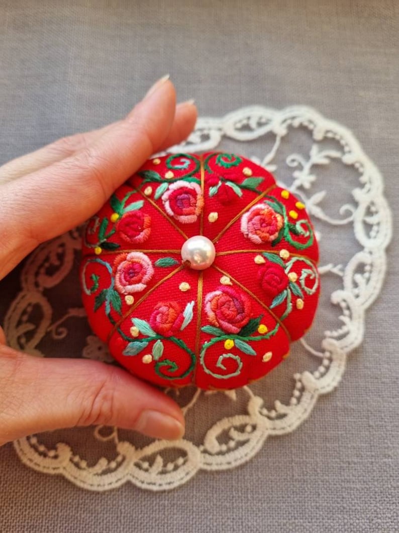 May include: A red pincushion with a pearl button in the center, embroidered with pink and red roses and green leaves.