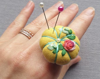 May include: A yellow fabric ring cushion featuring embroidered floral details in pink and green, accented with light blue thread segments and a central white bead. Two sewing pins with a white and a pink spherical head are inserted into the top.