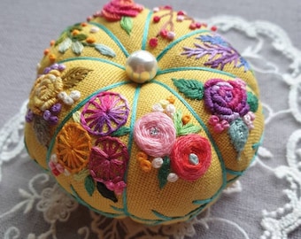 May include: A yellow fabric pincushion with a pearl button in the center. The pincushion is embroidered with colorful flowers and leaves.
