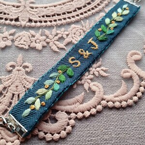 May include: A blue herringbone fabric bracelet featuring hand-embroidered green leaves, small yellow berries, and the gold-stitched letters S & J. The bracelet is finished with silver-toned metal clasps at both ends for a secure closure.