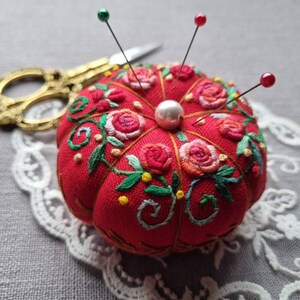 May include: A red pincushion with floral embroidery and a pearl button in the center. Four pins with colored heads are stuck into the pincushion.