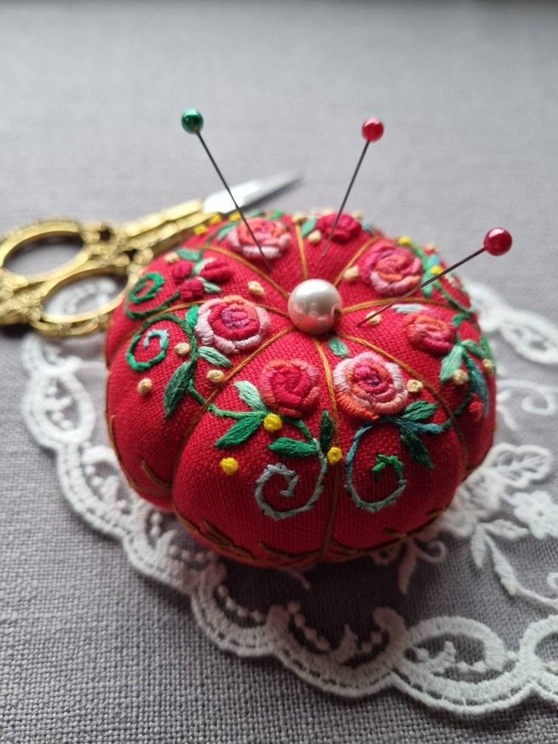 May include: A red pincushion with floral embroidery and a pearl button in the center. Four pins with colored heads are stuck into the pincushion.