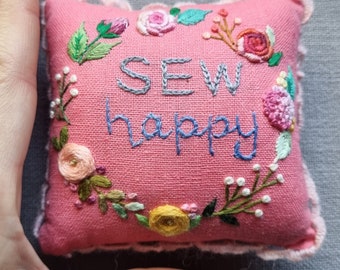 May include: A pink fabric pincushion with a white lace trim and embroidered flowers around the edge. The text "SEW happy" is embroidered in the center in gray and blue thread.