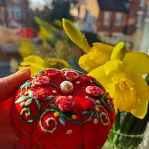 May include: A red pincushion with a pearl button in the center, embroidered with green leaves and pink flowers. The pincushion is held in a hand with a blurred background of yellow daffodils and a brick building.