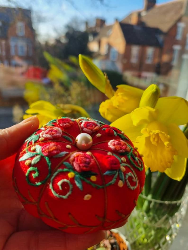 May include: A red pincushion with a pearl button in the center, embroidered with green leaves and pink flowers. The pincushion is held in a hand with a blurred background of yellow daffodils and a brick building.
