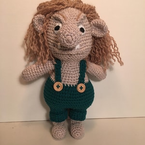 May include: A crocheted troll doll with brown hair, a gray body, and green overalls with brown buttons. The troll has a grumpy expression with a furrowed brow and a toothy grin.