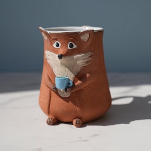 Peut inclure: Une tasse en céramique fantaisiste en forme de renard, tenant une petite tasse bleue. La tasse est principalement marron avec des accents blancs et noirs, représentant un visage et des oreilles de renard. La tasse est posée sur une surface blanche.