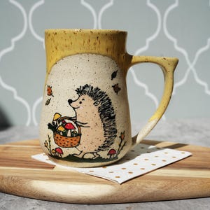 May include: A ceramic mug with a whimsical hedgehog design. The mug features a yellow rim and handle, with a beige body adorned with a hand-drawn hedgehog carrying a basket of mushrooms. Autumn leaves and mushrooms are scattered around the hedgehog.