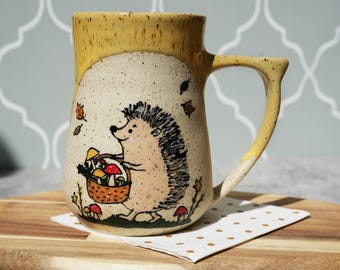 May include: A ceramic mug with a whimsical hedgehog design. The mug features a yellow rim and handle, with a beige body adorned with a hand-drawn hedgehog carrying a basket of mushrooms. Autumn leaves and mushrooms are scattered around the hedgehog.