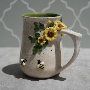 Op de afbeelding: Handgemaakte keramische mok met een gespikkelde beige afwerking. De mok heeft gele zonnebloem- en groene bladversieringen, samen met twee kleine bijenbeeldjes. De rand is groen en het handvat is wit.