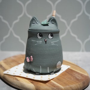 May include: A grey ceramic mug shaped like a cat with a lid. The cat features white painted whiskers, a pink nose, and a small pink butterfly on its side. A tiny green leaf sits on the lid, and a wooden stirrer is visible protruding from the top.