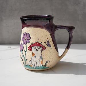 May include: A ceramic mug with a purple and cream glaze. The mug features a hand-painted illustration of a cat wearing a red mushroom hat, surrounded by flowers and a butterfly.