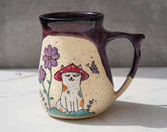 May include: A ceramic mug with a purple and cream glaze. The mug features a hand-painted illustration of a cat wearing a red mushroom hat, surrounded by flowers and a butterfly.