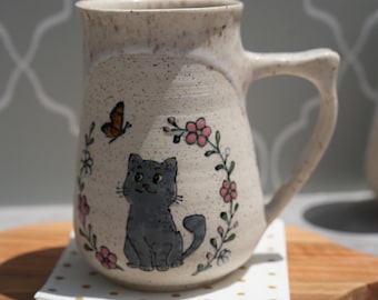 May include: A speckled off-white ceramic mug featuring a hand-painted illustration of a grey cat sitting in the centre. The cat is surrounded by a floral wreath with pink flowers and green stems, with a small orange butterfly above the cat.