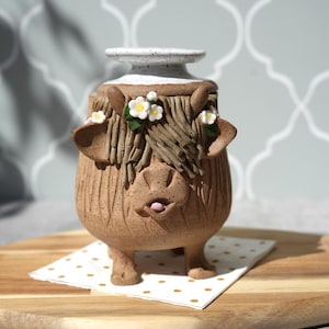 Op de afbeelding: Een grillige keramische vaas in de vorm van een Schotse Hooglandkoe. De bruine vaas heeft een witte rand, getextureerde vachtdetails en kleine witte bloemen. De koe heeft een speelse uitdrukking met haar tong uit. Het staat op een servet met stippen.
