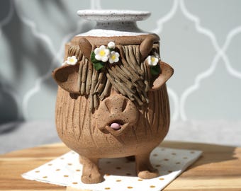May include: A whimsical ceramic vase shaped like a Highland cow. The brown vase features a white rim, textured fur details, and small white flowers. The cow has a playful expression with its tongue sticking out. It sits on a polka-dot napkin.