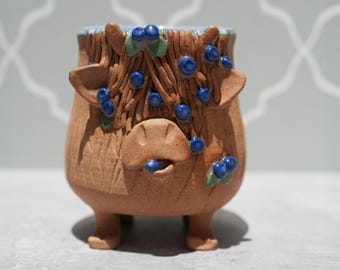 May include: A whimsical, handmade ceramic planter in the shape of a Highland cow. The brown stoneware pot features a textured surface, small legs, and is adorned with blue blueberry accents and green leaves. The rim is painted light blue.