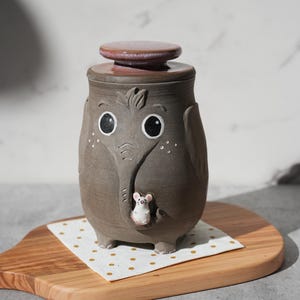 May include: A whimsical ceramic jar shaped like an elephant, with a lid. The jar is gray with white accents and a small mouse figurine. The lid is a reddish-brown color. The jar is sitting on a wooden cutting board with a white and gold polka dot napkin.