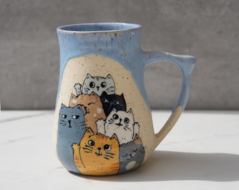 May include: A blue ceramic mug with a white glaze and a handle. The mug features a design of six cats stacked on top of each other, with their faces peeking out from behind each other.