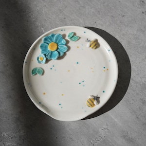 Op de afbeelding: Witte keramische plaat versierd met een blauwe bloem, groene blaadjes en twee geel-zwarte bijen. De plaat heeft een kleine inkeping en is versierd met blauwe en gele stippen.
