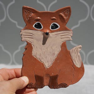 May include: A ceramic fox figurine in shades of brown and cream. The fox has large black eyes, a cream-colored chest, and a detailed tail. The figurine is held in a hand, showcasing its size and shape. The background is a light gray with a repeating pattern.