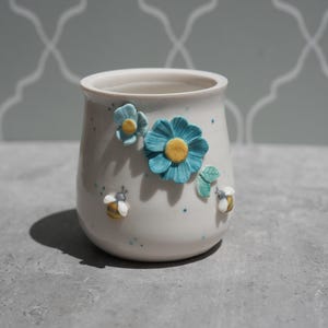 Puede incluir: Una maceta de cerámica blanca con adornos de flores azules y abejas. La maceta tiene forma cilíndrica con una parte superior ligeramente ensanchada. Las flores tienen centros amarillos y las abejas rayas amarillas y blancas.