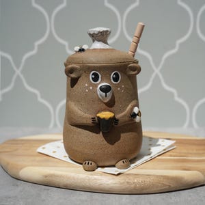 May include: A speckled brown ceramic honey pot shaped like a bear, featuring a lid with a white handle, a small bee figurine, and a wooden honey dipper. The bear holds a small honey pot, and a second bee is attached to its side, all in a matte finish.